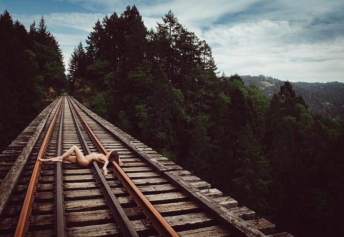 Nude brunette woman lying on her back across the wooden trestle train tracks with her left arm stretched to the side and left knee bent upwards gazing at the tracks leading into the forest.