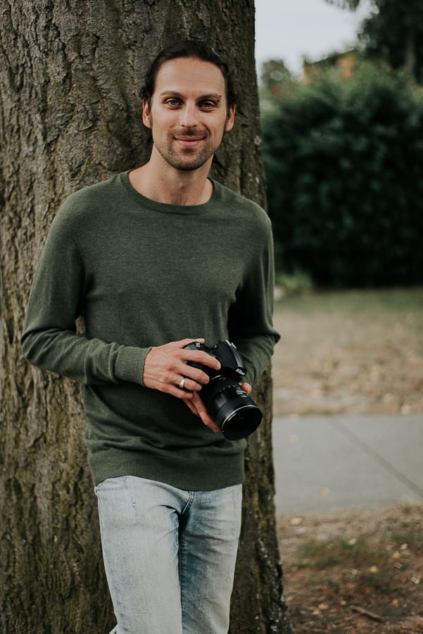 man with brown hair and green sweater leaning up against a tree holding a camera
